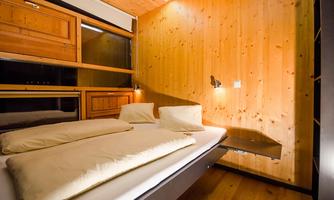 A modern bedroom with paneled wooden walls. The bed is equipped with two pillows and blankets, and there is a small table against the wall.