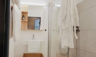 A modern bathroom with a glass shower and a bright, minimalist design. Two white bathrobes hang on a towel rack.