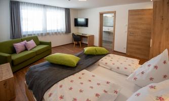 A modern hotel room with a double bed and fresh floral patterns. In the background, there is a couch, a table, and a television.