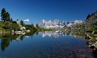 A clear lake reflects majestic mountains and a blue sky. Surrounded by lush greenery and rocks, the landscape offers a peaceful atmosphere.