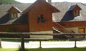 A traditional wooden house with a beautiful balcony and flowering plants. In the background, green hills and a blue sky can be seen.