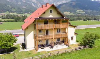 A cozy house with several balconies, surrounded by green meadows and mountains. In the background, a road runs by.