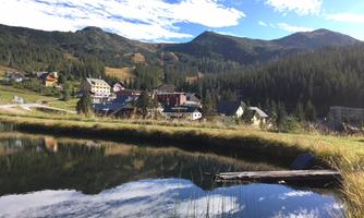 A peaceful landscape with a clear pond and mountains in the background. Houses line the shoreline, surrounded by trees and meadows.