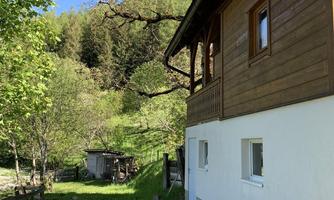 An idyllic house surrounded by trees and green meadow. In the background, mountains and a clear sky can be seen.