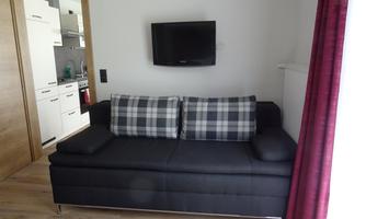 A modern living room with a black sofa and checkered cushions. In the background, an open kitchen with white cabinets can be seen.