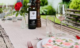 A beautifully set table outdoors with a wine bottle, glasses, and a red rose. The table is decorated with an inviting tablecloth and dishware.