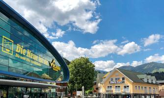 A modern building facade with large windows and yellow lettering. In the background, mountains and a mix of shops and restaurants can be seen.