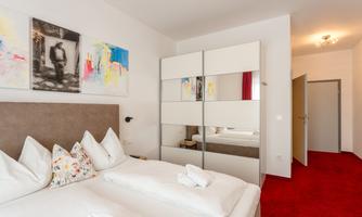A modern bedroom with a double bed and a large mirror wardrobe. The walls are painted light, and the decor is simple and colorful.