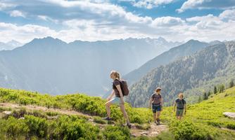 Three hikers explore a green mountain landscape. The mountain backdrop and the blue sky create a relaxed atmosphere.