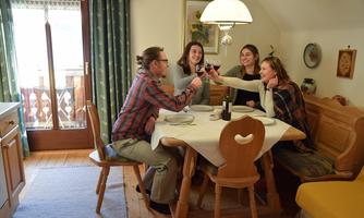 A cozy gathering at the table with four people toasting with wine. In the background, a bright room with a window and wooden details is visible.