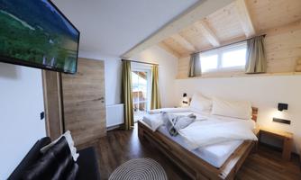 A cozy bedroom with a large double bed and wooden details. There is a television on the wall and windows with curtains that let in natural light.