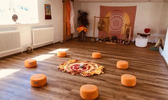 A quiet room with wooden flooring and orange cushions arranged in a circle. In the background, colorful wall hangings and meditation materials can be seen.