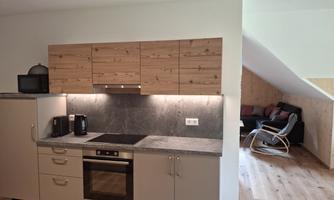 A modern kitchen with wooden shelves and a gray workspace. In the background, there is a cozy living area.