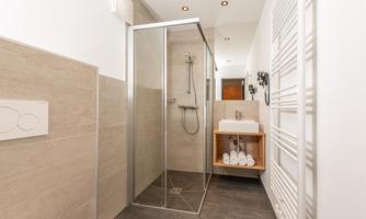 A modern bathroom with a clear glass shower and a small sink. Towels are neatly stacked on a wooden shelf.