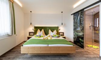 A modern bedroom with a large bed and green bedding. The room is bright and has a glass shower to the side.