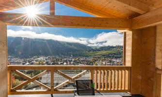 A sunny view from a balcony of a picturesque town and surrounding mountains. The clouds float gently over the hills as the sunlight shines through the wooden building.