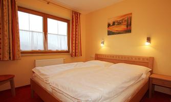 A cozy bedroom with a double bed and red carpet. The walls are painted in a warm shade and a window provides natural light.