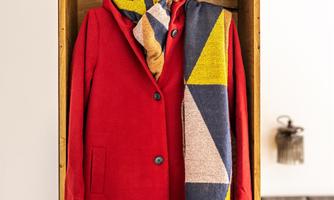 A red coat hangs in a wooden shelf, accompanied by a colorful scarf and a ski mask. The background shows a cozy living environment.