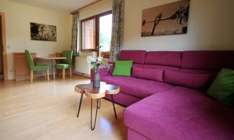 A cozy living room with a purple couch, a small wooden table, and a vase with flowers. In the background, there is a dining area and windows with a view to the outside.