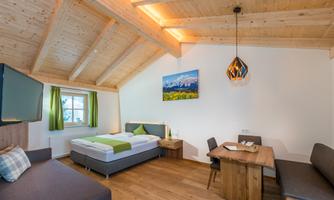 A modern room with a wooden beam ceiling, a double bed, and a cozy seating area. There is a window with green curtains and a table for meals.
