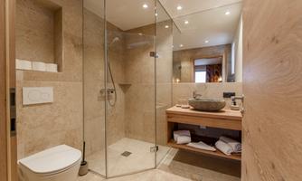 A modern bathroom with a glass shower and a wooden vanity. The walls are covered in beige stone, and there is a stylish toilet.