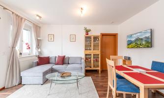 A modern living room with a grey sofa and a glass table. A dining area with wooden chairs and a colorful table is also present.