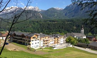 A picturesque mountain landscape with snow-capped peaks and green meadows. In the foreground, modern residential buildings stand next to a small village.