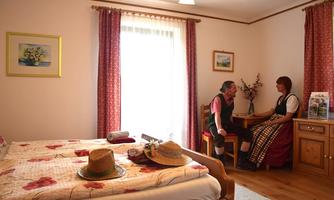 A cozy room with a double bed, a wooden dresser, and a small table. A couple in traditional clothing is sitting at the table and having a conversation.