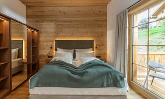 A modern bedroom with a comfortable double bed and a warm wood design. Large windows offer a view outside and provide plenty of daylight.