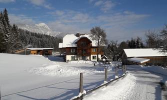 A cozy wooden house in a snowy landscape. Surrounded by trees and mountains under a clear sky.
