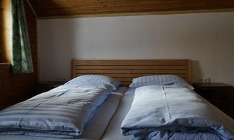 A cozy bedroom with a double bed and blue bedspreads. Wooden paneling and a window with green curtains create a warm atmosphere.