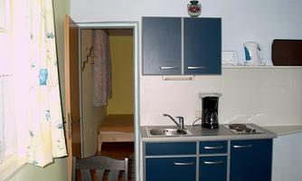 A small kitchen with blue cabinets and a table. In the background, an open room with a bed can be seen.