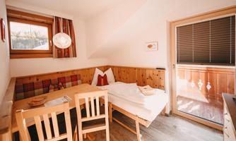 A cozy room with wooden paneling and a small table with chairs. There is a window with a view of a balcony and a decorative ceiling lamp.