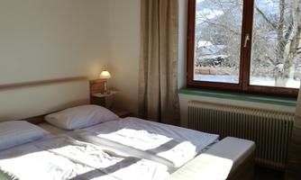 A bright bedroom with a double bed and a window offering a view of a snow-covered landscape. The soft curtains and the bedside lamp create a cozy atmosphere.