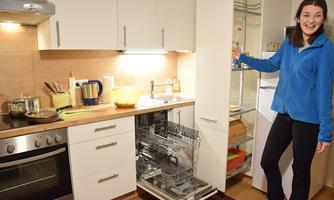 A modern kitchen with an open dishwasher and a refrigerator door. A cheerful person proudly shows off the clean surroundings.