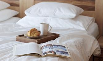 A cozy bed with white sheets and pillows. On a tray are a croissant, a cup of coffee, and an open book.