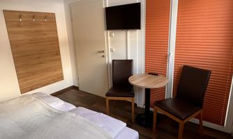 A cozy room with a bed, two chairs, and a small table. The windows have orange blinds and there is a television on the wall.