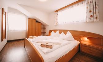 A cozy bedroom with a wooden bed and three pillows. The large windows provide plenty of daylight.