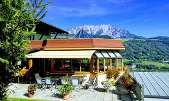 A charming building with large windows and a terrace, surrounded by green meadows. In the background, majestic mountains can be seen under a clear blue sky.