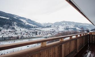 A view from the balcony of a snowy mountain landscape. In the foreground, the wooden railing of the balcony is visible.