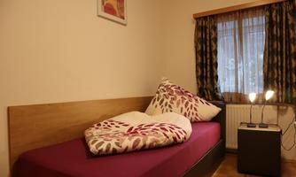 A simple bedroom with a bed adorned with a colorful bedspread and pillows. A small nightstand with a lamp is next to the bed, and a window with curtains lets soft light in.