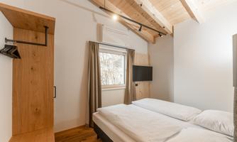 A cozy bedroom with a wooden ceiling, two beds, and a window. There is also a wardrobe and a television on the wall.