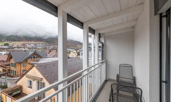 A balcony with two chairs and a view of a picturesque town. In the background, mountains and clouds are visible.