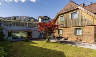 A charming house with wooden cladding and a beautiful terrace. In the background, mountains and a blue sky can be seen.