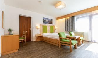 A modern hotel room with a double bed, a wooden dresser, and two armchairs. Large windows let in plenty of daylight and create a friendly atmosphere.