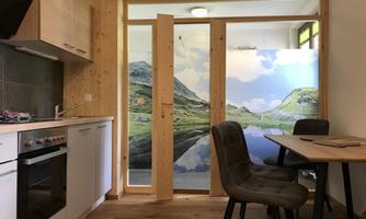 A modern kitchenette with wood elements and a dining area. In the background, there are doors with a picturesque landscape view.