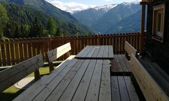 A wooden terrace with several tables and benches. Impressive mountains and a clear sky are visible in the background.