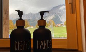 Two elegant soap dispensers, one for dishes and one for hands, are placed on a wooden coaster. In the background, the mountains and a cloudy sky can be seen.