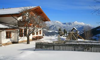 An idyllic winter landscape featuring a cozy house and snow-covered mountains in the background. In the foreground, there is a playground surrounded by a wooden fence and snow-covered trees.