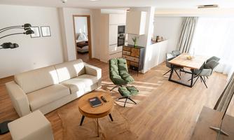 A modern living room with a comfortable white sofa and a round wooden table. The room has large windows that let in plenty of light.
